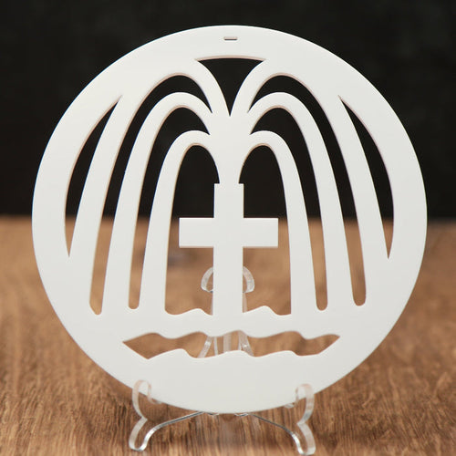 A white circular Christmas ornament with a cross in the center and fountains gushing from the cross, displayed on a clear stand on a wooden surface against a dark background.