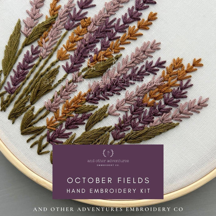 Close-up of a hand embroidery kit in progress, showing embroidered lavender stems in purple, mauve, brown, and olive green on white fabric inside a wooden hoop. A purple label reads 'October Fields Hand Embroidery Kit' with the brand 'And Other Adventures Embroidery Co' visible.